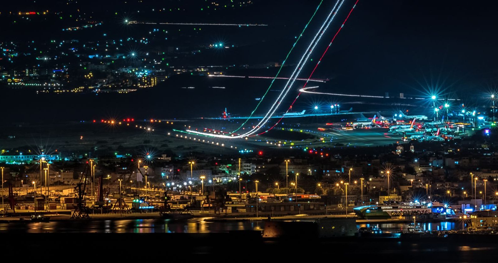 Vista aérea noturna de uma cidade com aeroporto e área portuária iluminados.