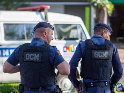 Dois guardas municipais uniformizados patrulham uma rua urbana.