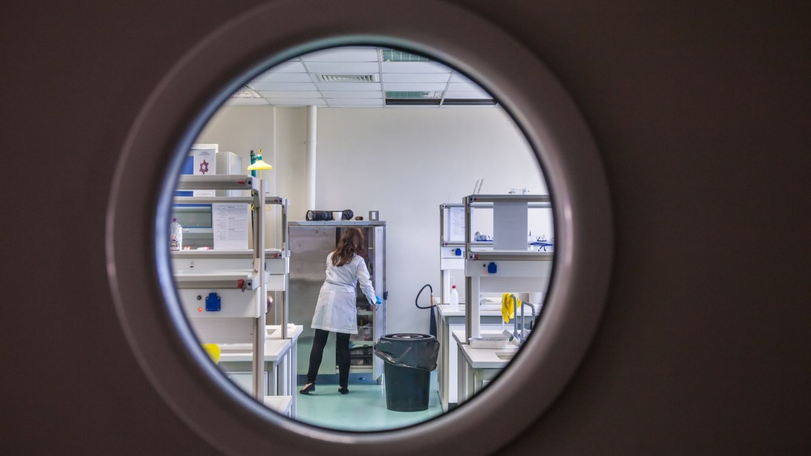 Cientista usando jaleco trabalha em um laboratório moderno, observada por uma janela circular.
