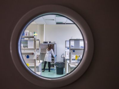 Cientista usando jaleco trabalha em um laboratório moderno, observada por uma janela circular.