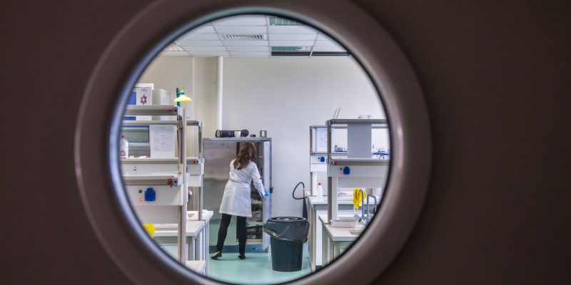 Cientista usando jaleco trabalha em um laboratório moderno, observada por uma janela circular.